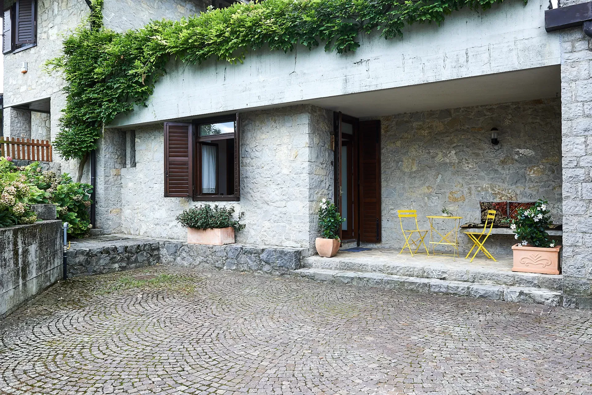 Facciata in pietra di una casa con patio, finestre con persiane e tavolino giallo all’aperto