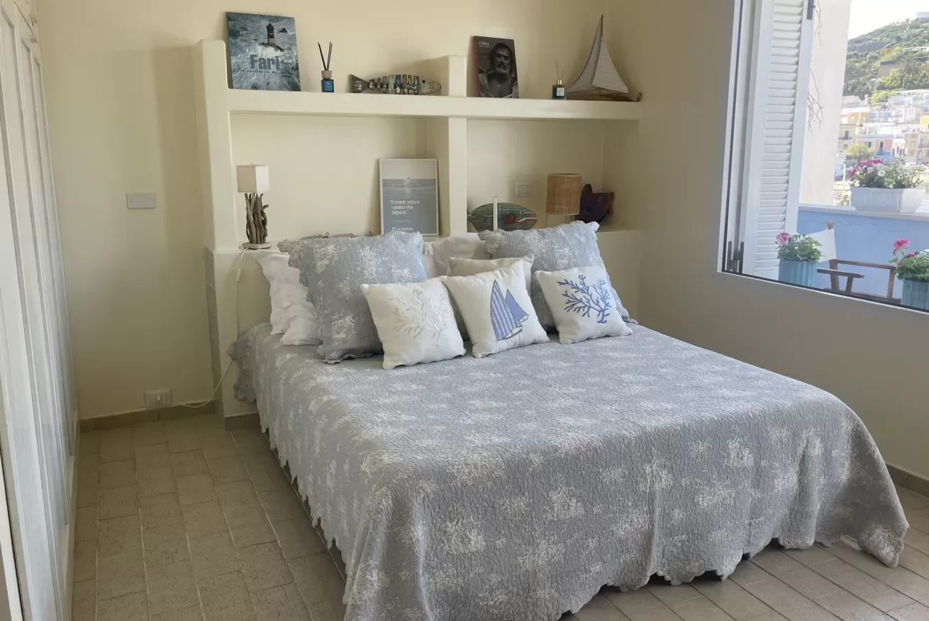 Camera da letto luminosa con letto matrimoniale, cuscini decorativi e finestra aperta con vista sul mare
