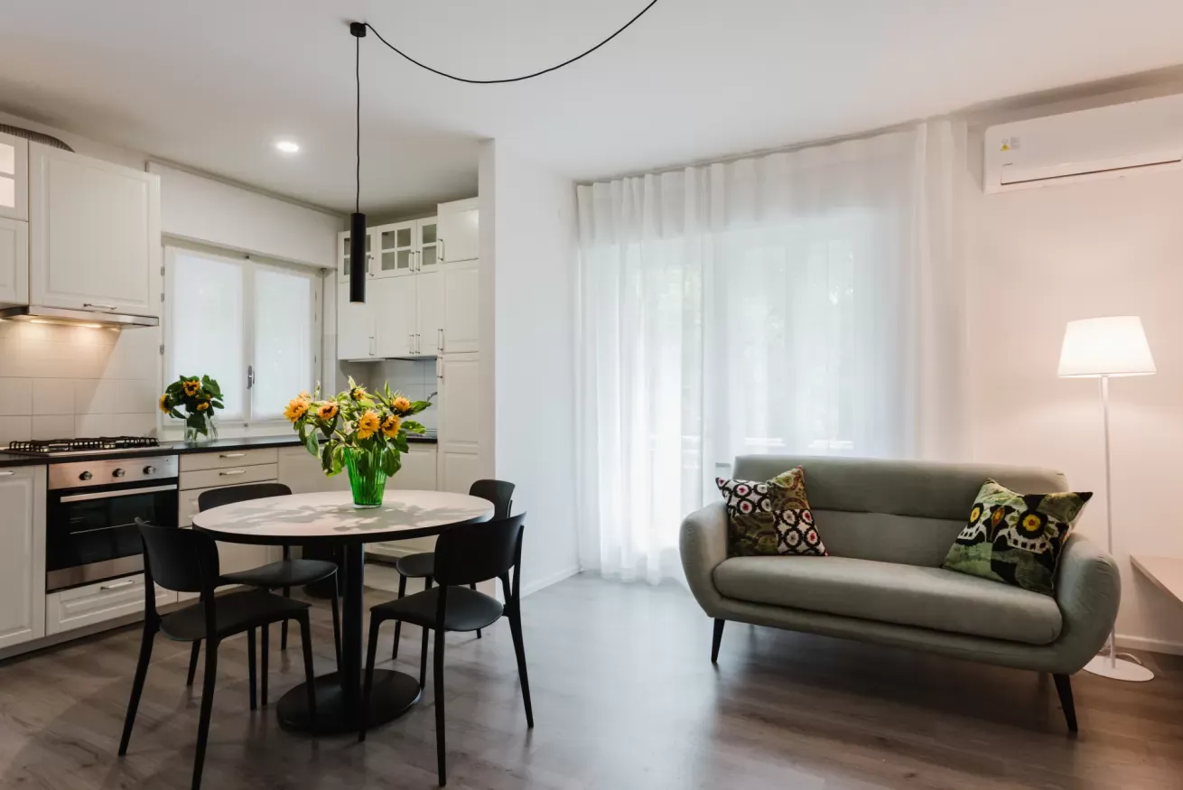 Soggiorno open space con cucina bianca, tavolo rotondo, divano grigio e grande finestra con tende bianche
