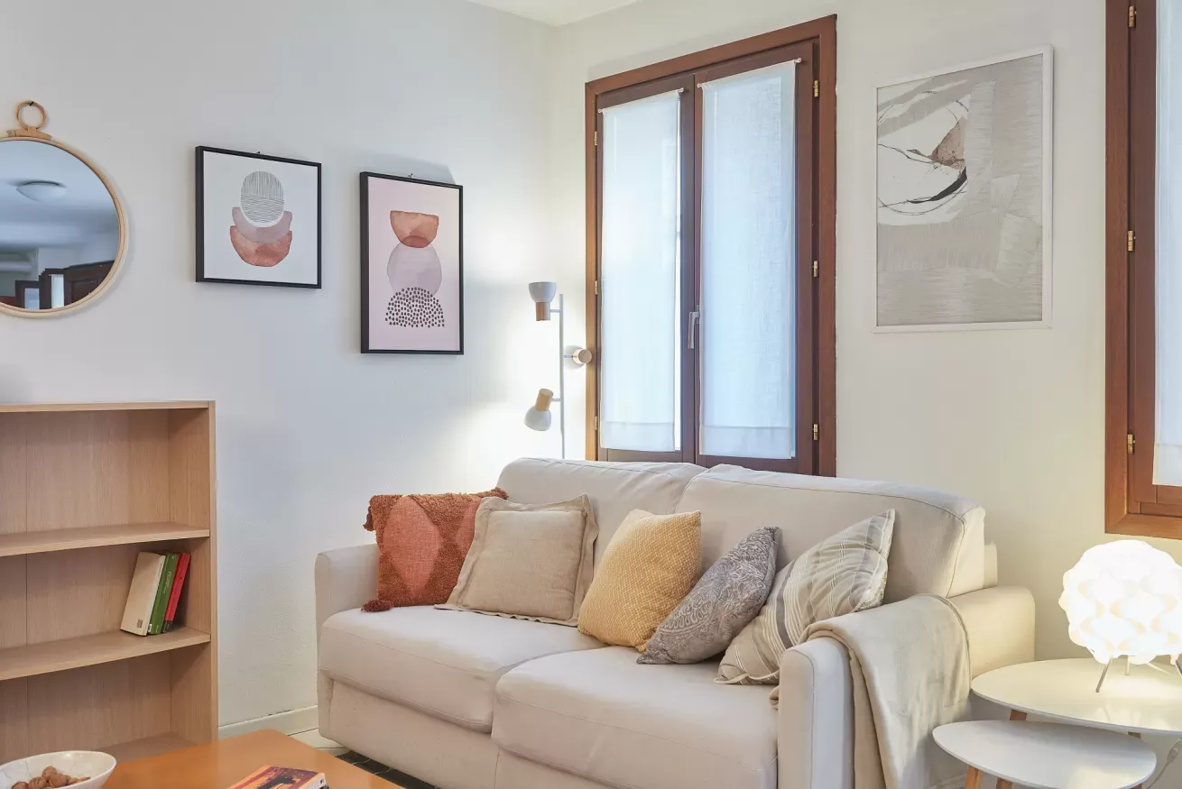 Salotto luminoso con divano chiaro, cuscini decorativi, quadri alle pareti e finestre in legno
