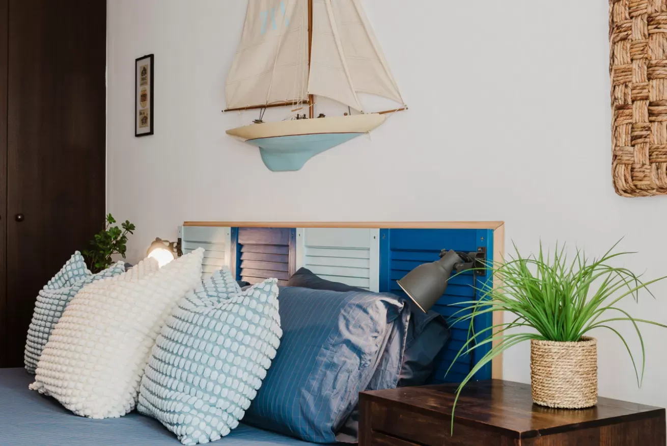 Camera da letto con letto blu, cuscini decorativi, lampada, pianta in vaso e modellino di barca a vela sulla parete