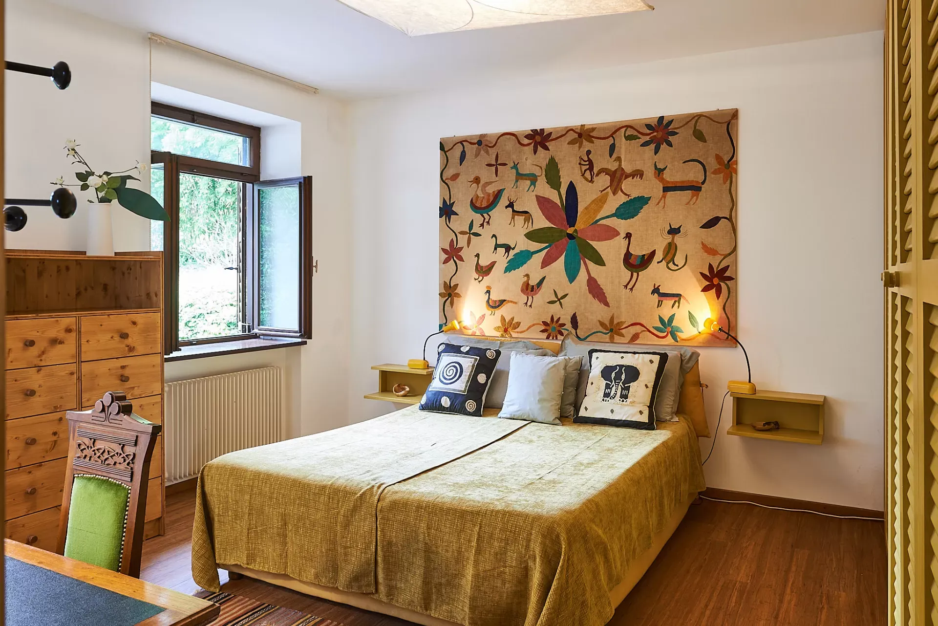 Camera da letto luminosa con letto matrimoniale, finestra aperta e arazzo decorativo sopra la testiera