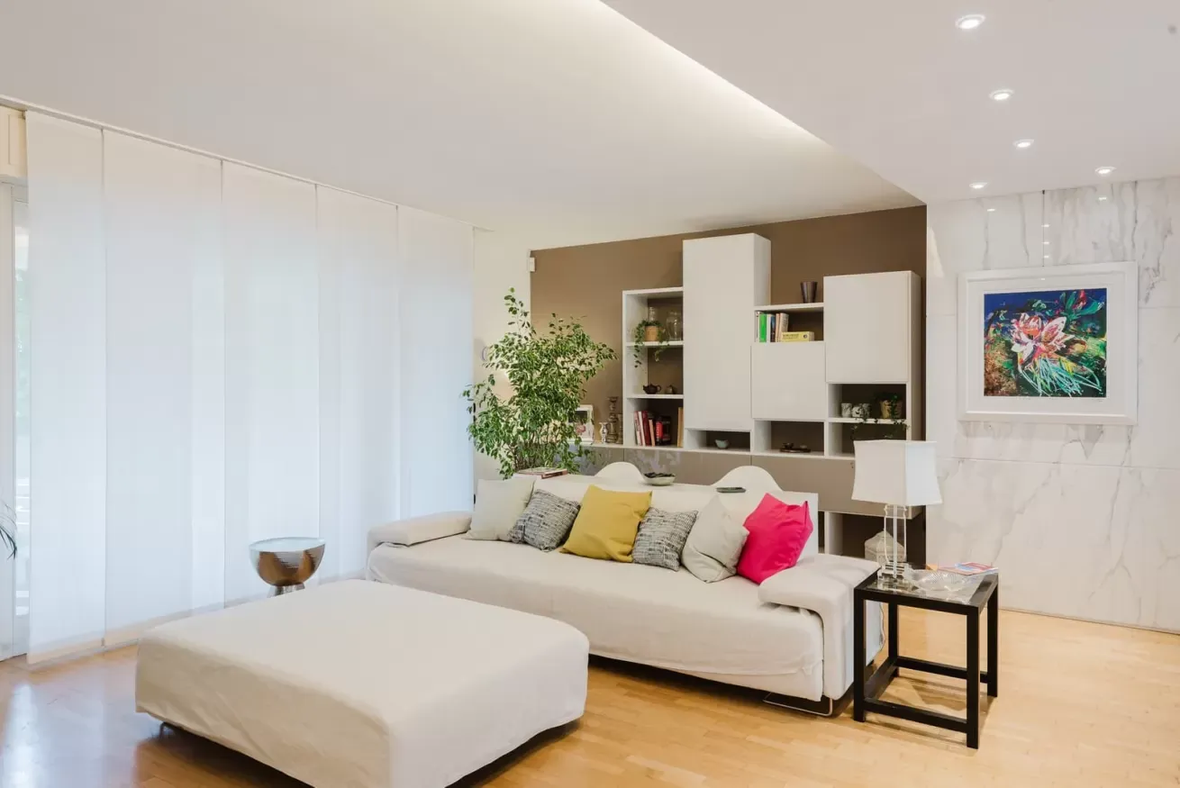 Soggiorno moderno e luminoso con divano chiaro, pouf, scaffalature a muro e grande finestra con tende bianche