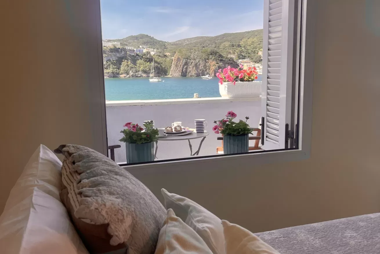 Vista sul mare da una finestra, con balcone fiorito e barca all’ancora in lontananza