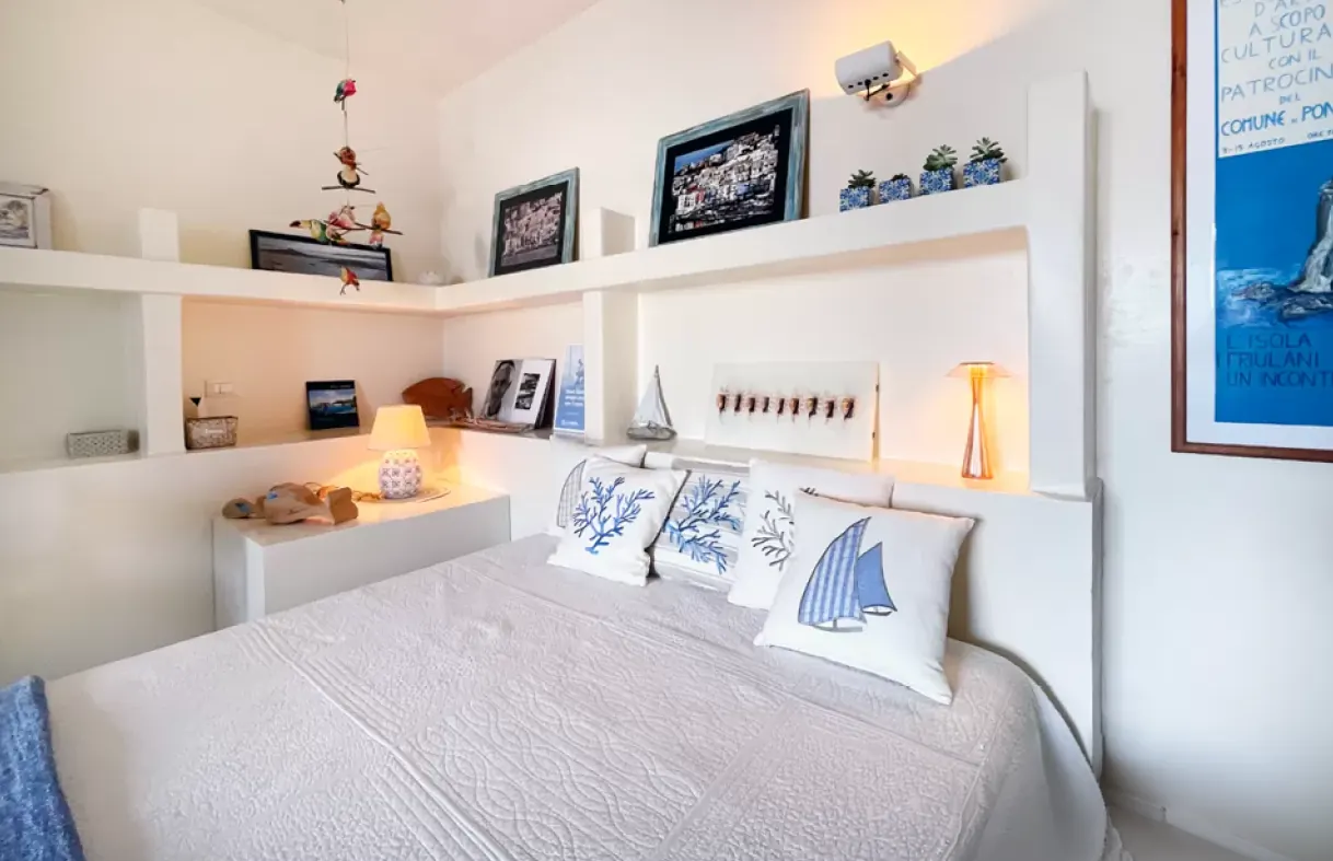 Camera da letto luminosa con letto matrimoniale, mensole bianche, lampade e cuscini decorativi a tema mare