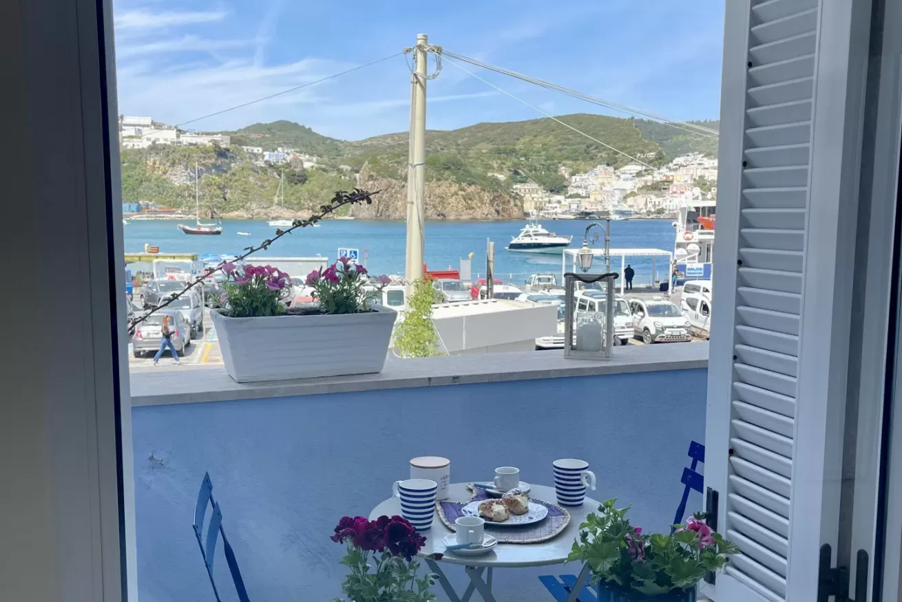 Vista su un porto con barche e colline, da una finestra con tavolino apparecchiato sul balcone