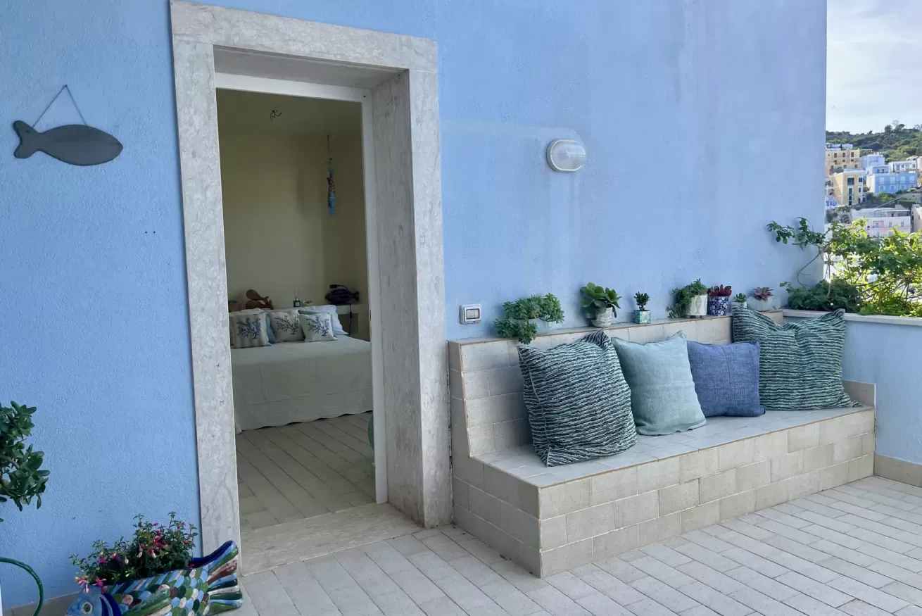 Terrazza con panca in muratura, cuscini e piante, accanto a un ingresso aperto verso una stanza