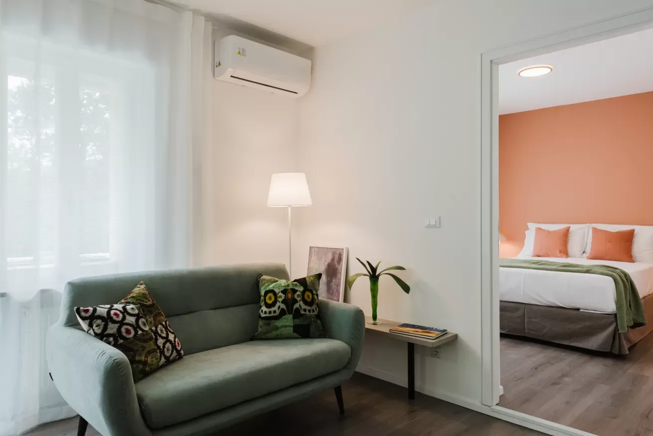 Salotto moderno con divano verde, lampada e vista su una camera da letto attraverso la porta aperta