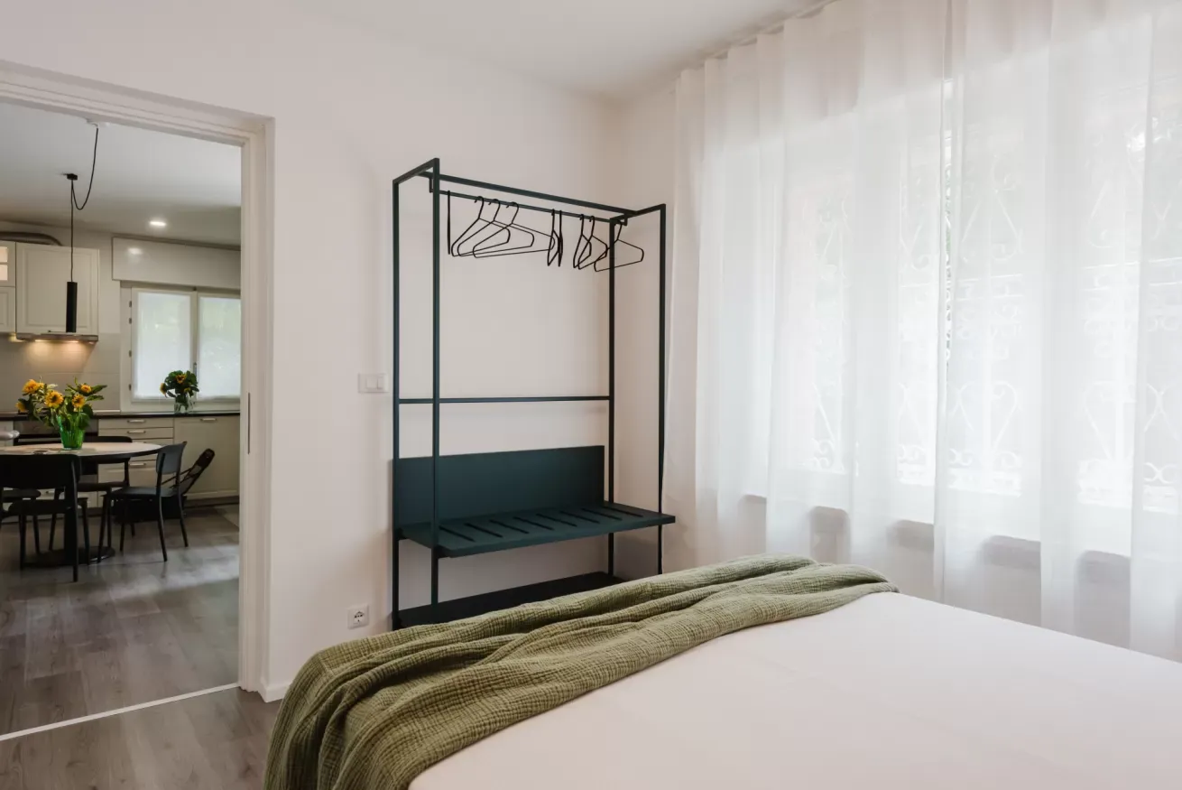 Camera da letto luminosa con letto, appendiabiti e finestra con tende bianche; si vede la cucina attraverso la porta aperta