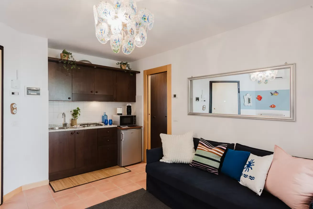 Soggiorno con angolo cottura, divano scuro, specchio a parete e lampadario decorativo