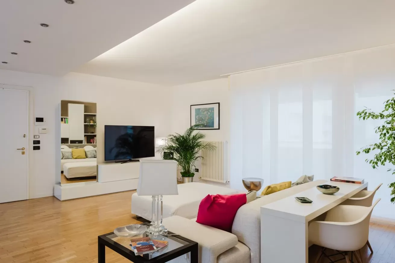 Soggiorno moderno e luminoso con divano bianco, TV, tavolo da pranzo e grandi finestre con tende bianche